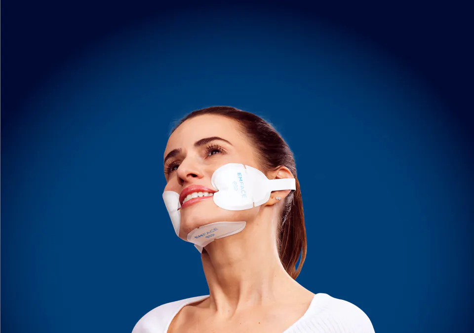 Smiling woman wearing EMFACE cheek and under-chin applicators, EMFACE face lift treatment at Square Mile Clinic, City of London